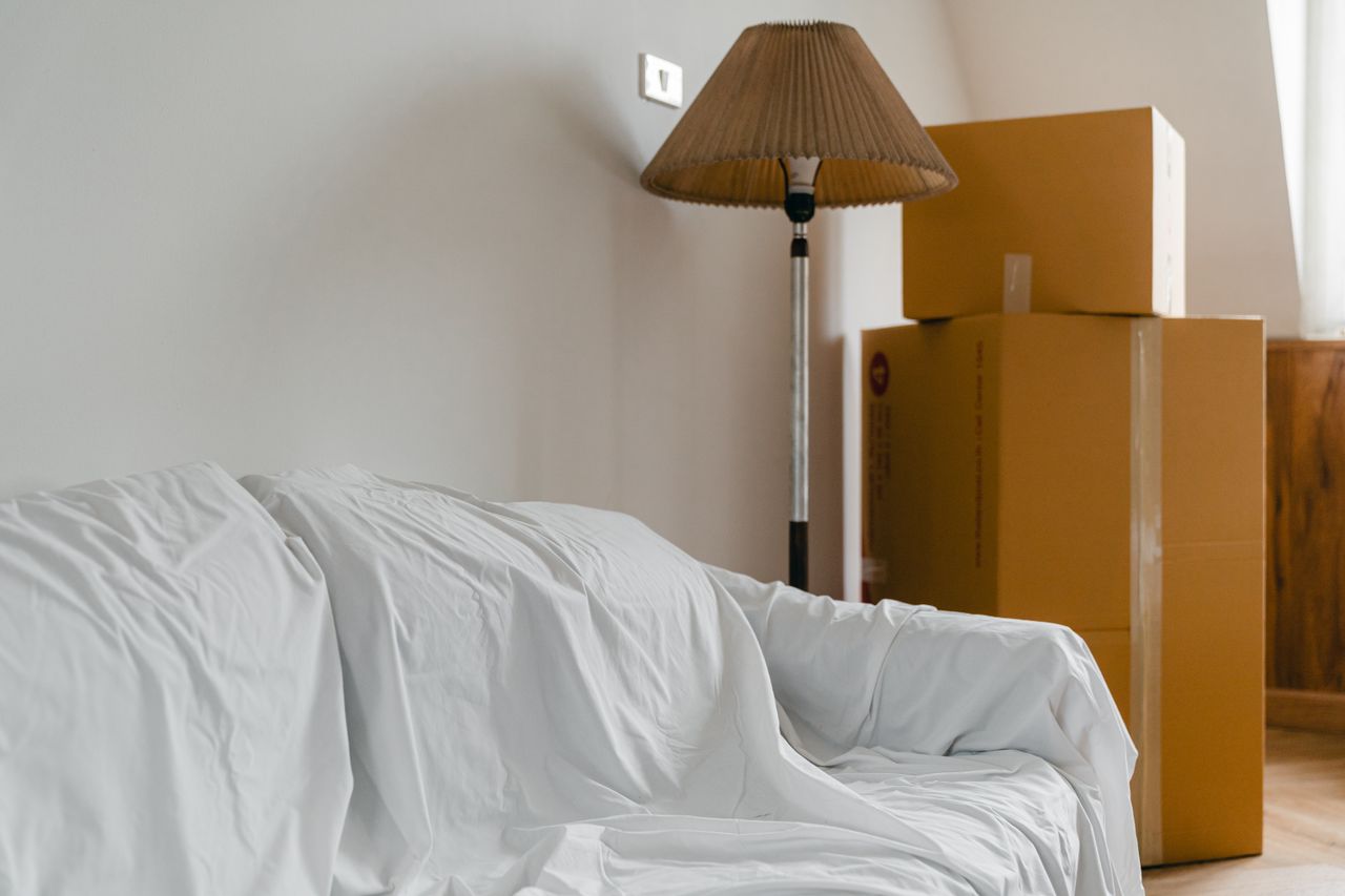 services-01 Covered with sheet sofa and floor lamp and packed cardboard boxes in light spacious room of new contemporary house